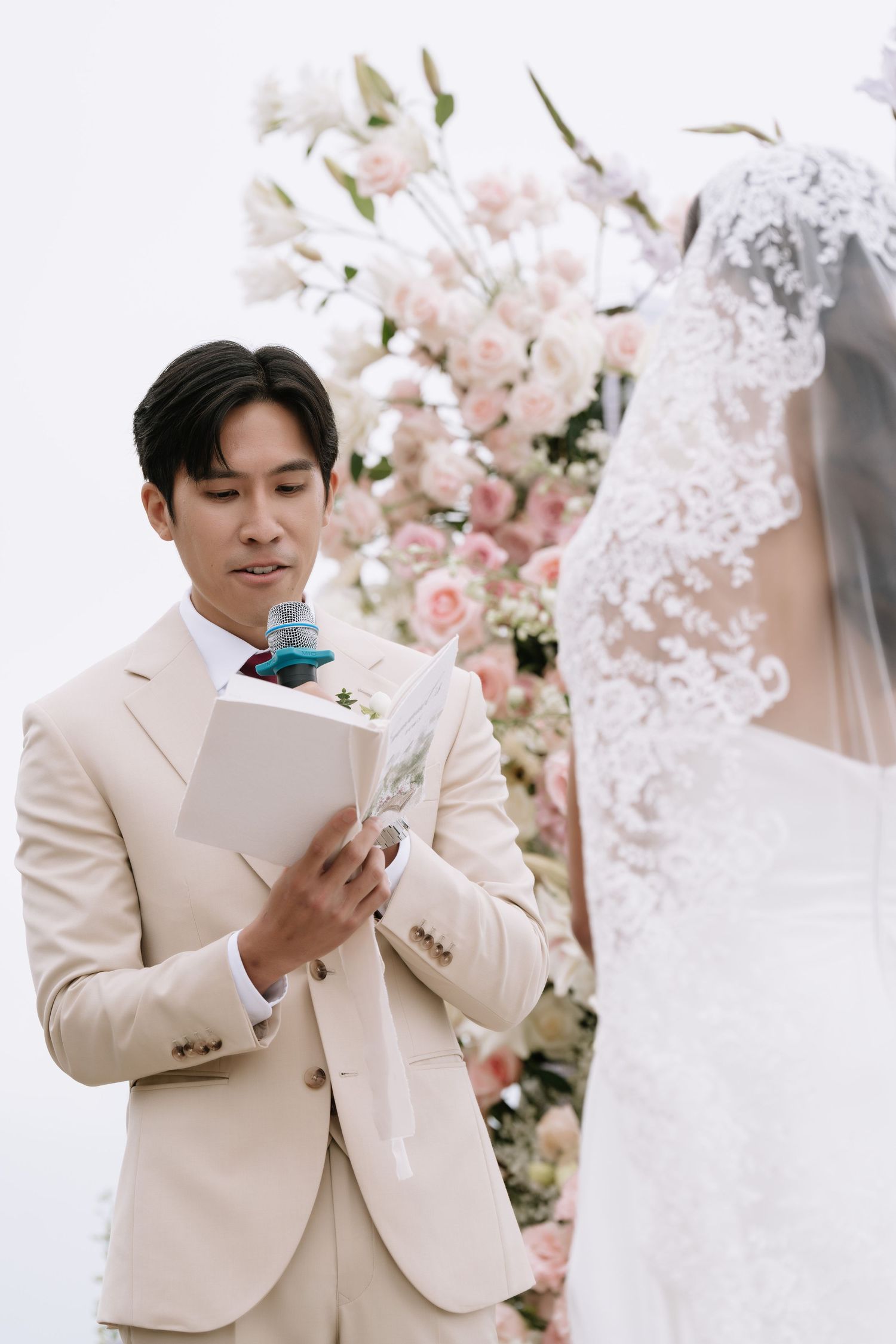 Handsome groom in a beige suit reads his wedding vows before his bride at a floral-decorated outdoor ceremony altar.