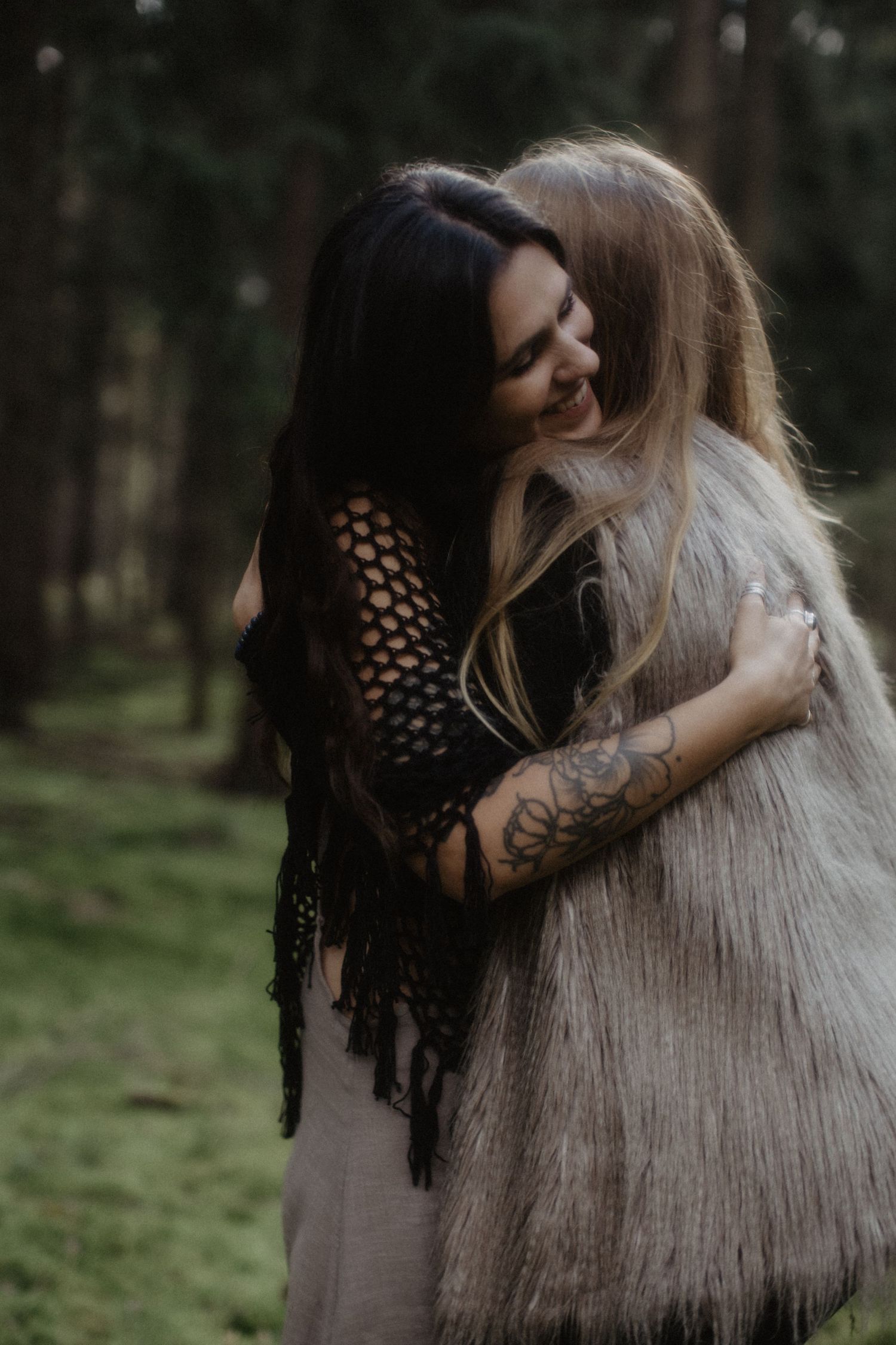 Frauen umarmen sich in einer stimmungsvollen Waldkulisse. Die schwarz-beige Kleidung erzeugt eine ätherische Atmosphäre.
