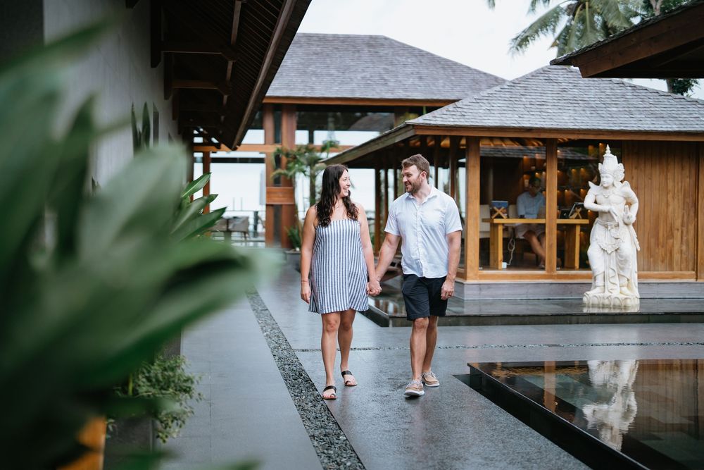A couple walks along a wooden boardwalk surrounded by tropical palm trees and lush greenery.