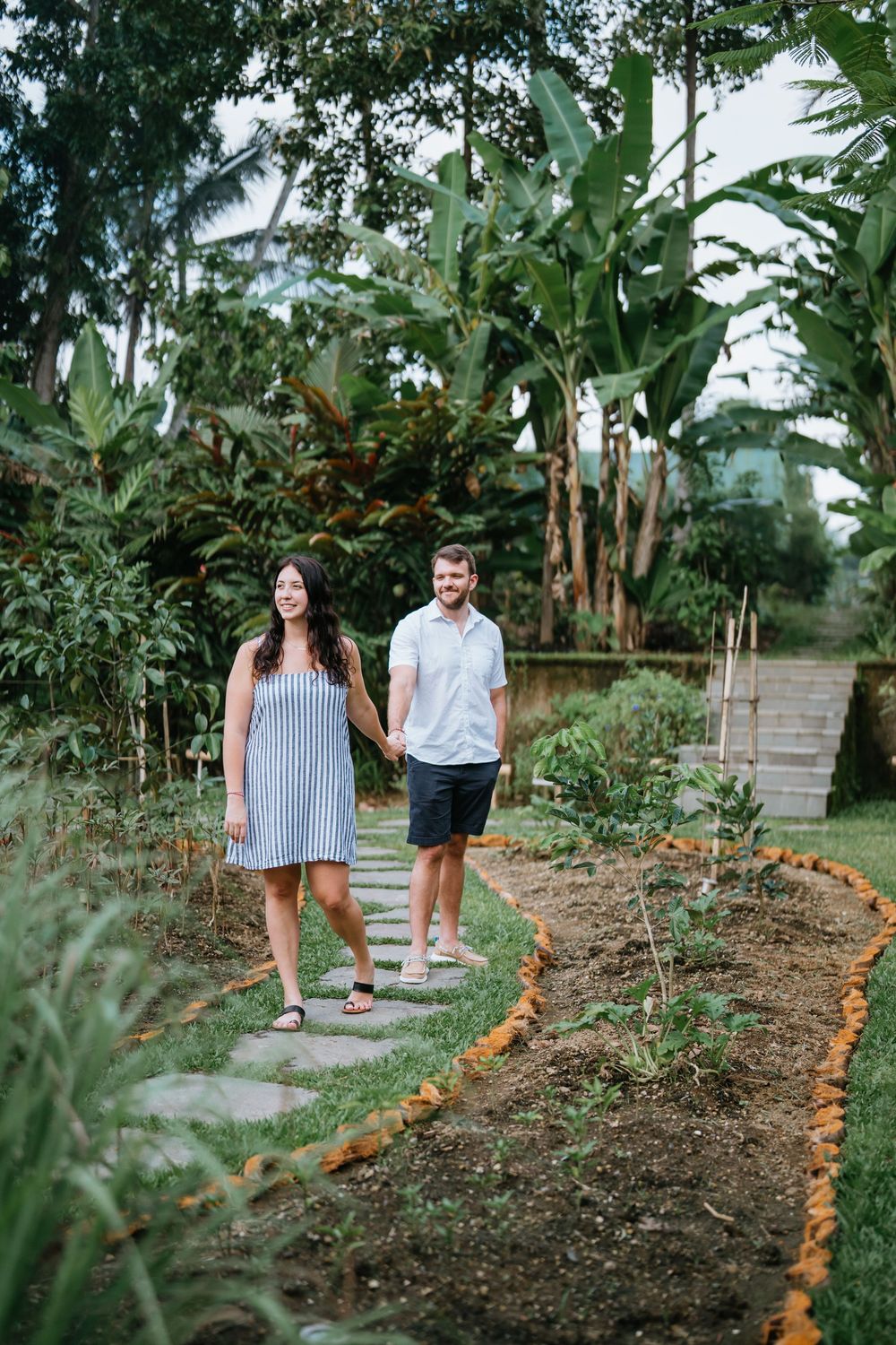 A couple follows a path lined with orange petals through a tropical garden setting.