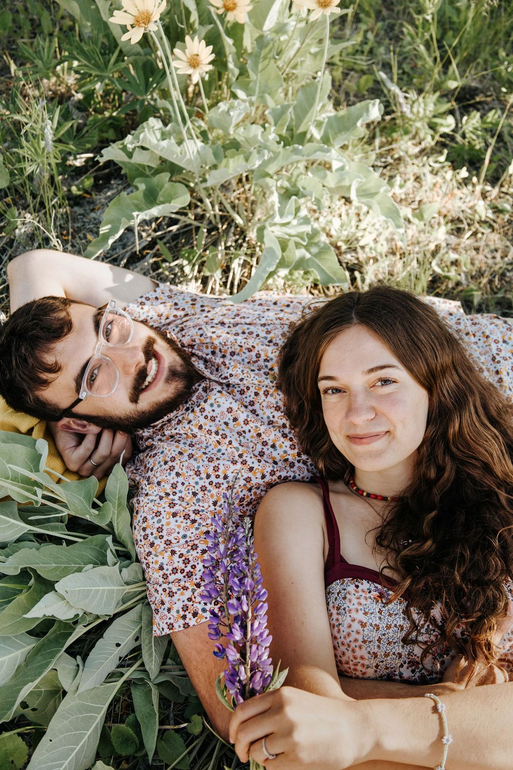 A couple lies in a meadow surrounded by blooming flowers and greenery.