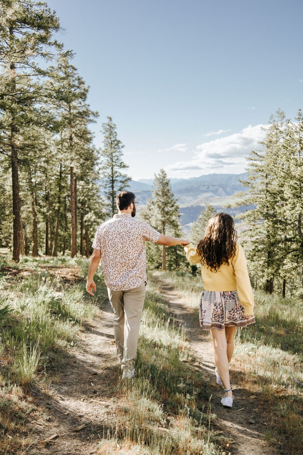 A couple runs joyfully down a mountain trail surrounded by pine trees.