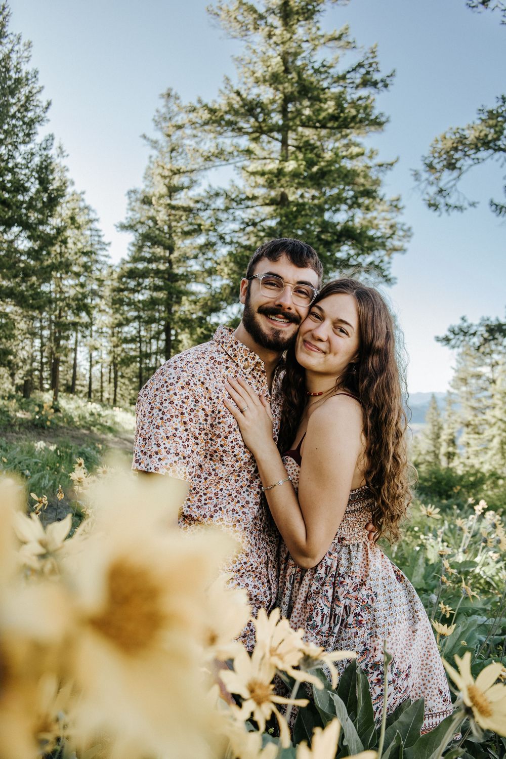 A couple shares an intimate moment among wildflowers in a mountain forest setting.