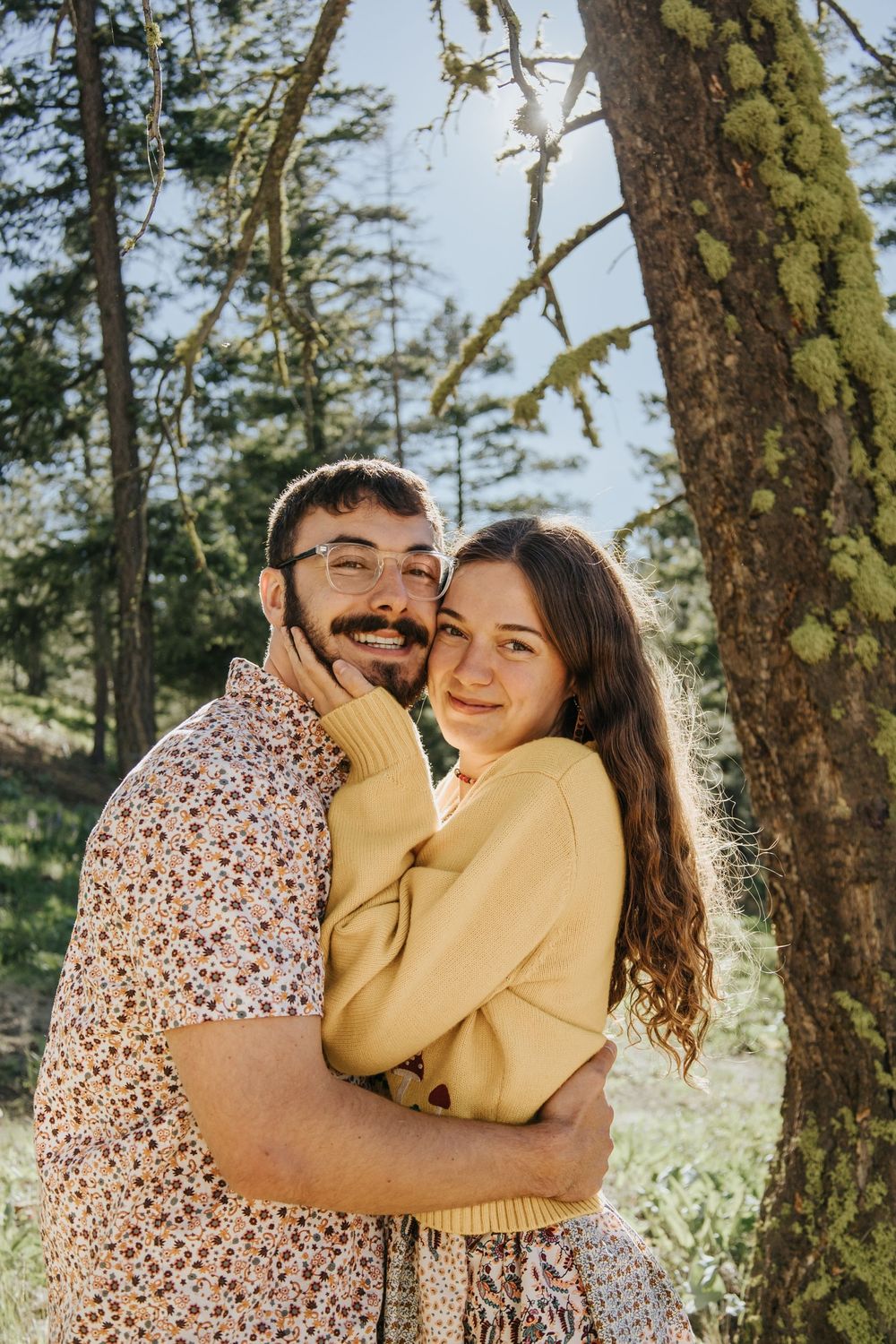 A romantic couple embraces under a mossy tree in a sunlit forest setting.