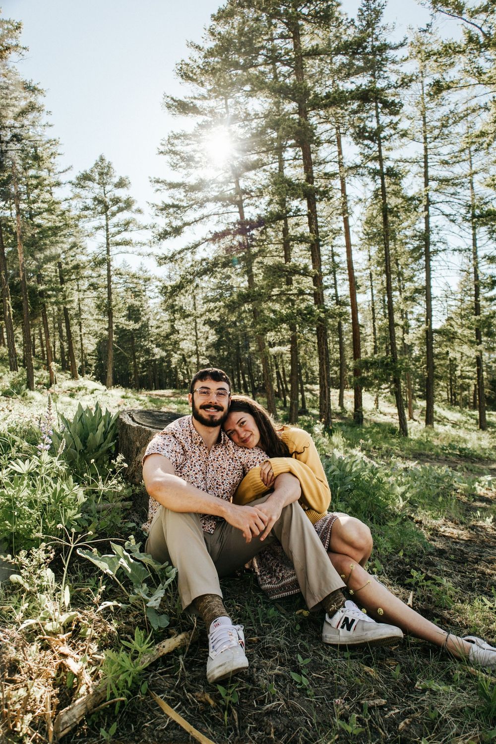 A couple relaxes together on a hillside surrounded by tall pine trees on a sunny afternoon.