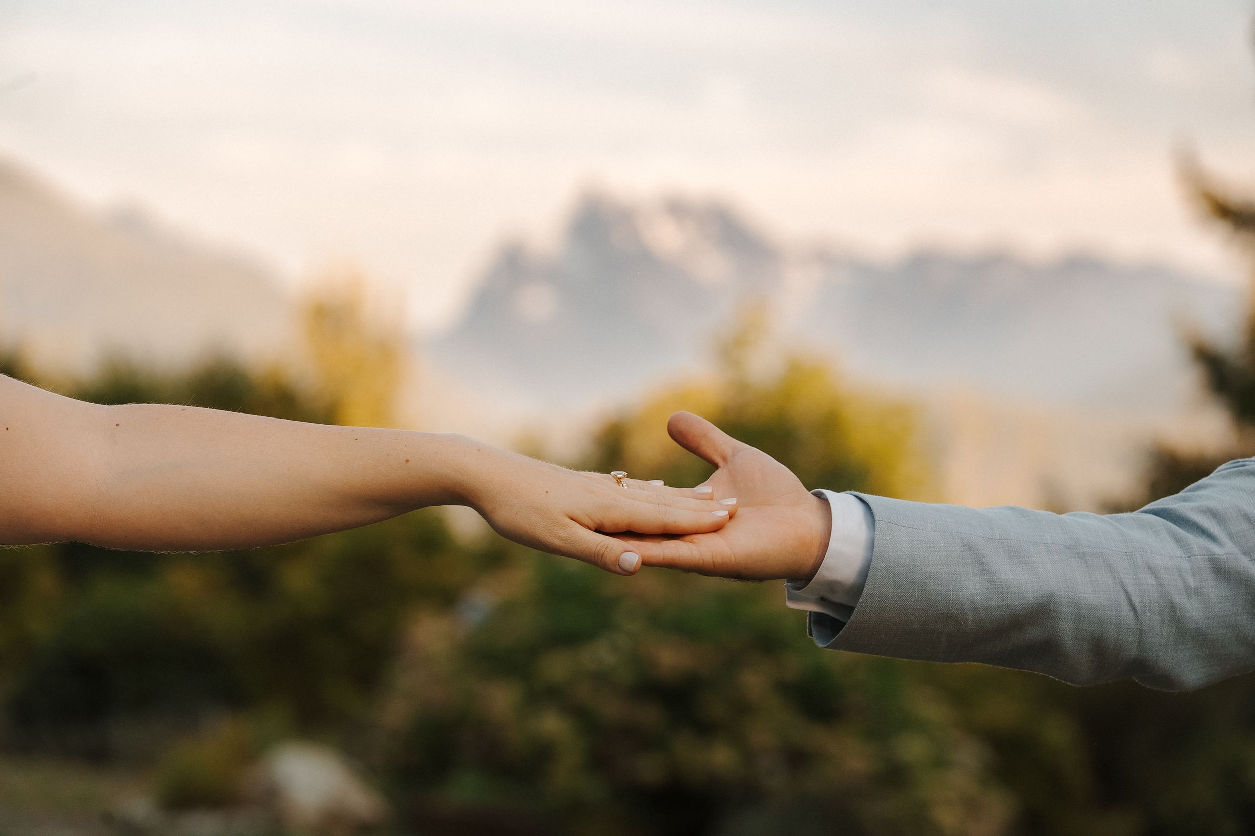 Two hands reaching out to touch each other in a tender moment against a blurred mountain landscape.