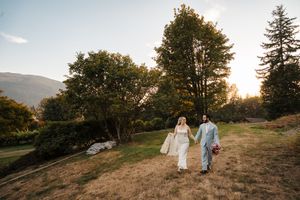 A couple walks hand in hand through a golden sunset meadow on a hilltop as warm light filters through trees.