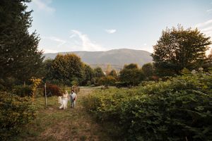 Two people walk a dirt path through a lush garden with mountains rising in the background on a sunny day.