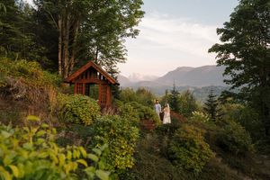 A rustic mountain cabin nestled among tall trees with scenic mountain peaks visible in the background at sunset.