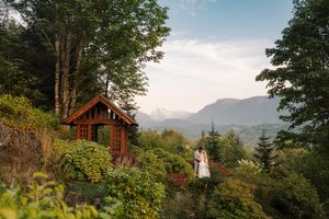 A log cabin nestled on a hillside among green trees and mountains with a scenic mountain vista in the background.
