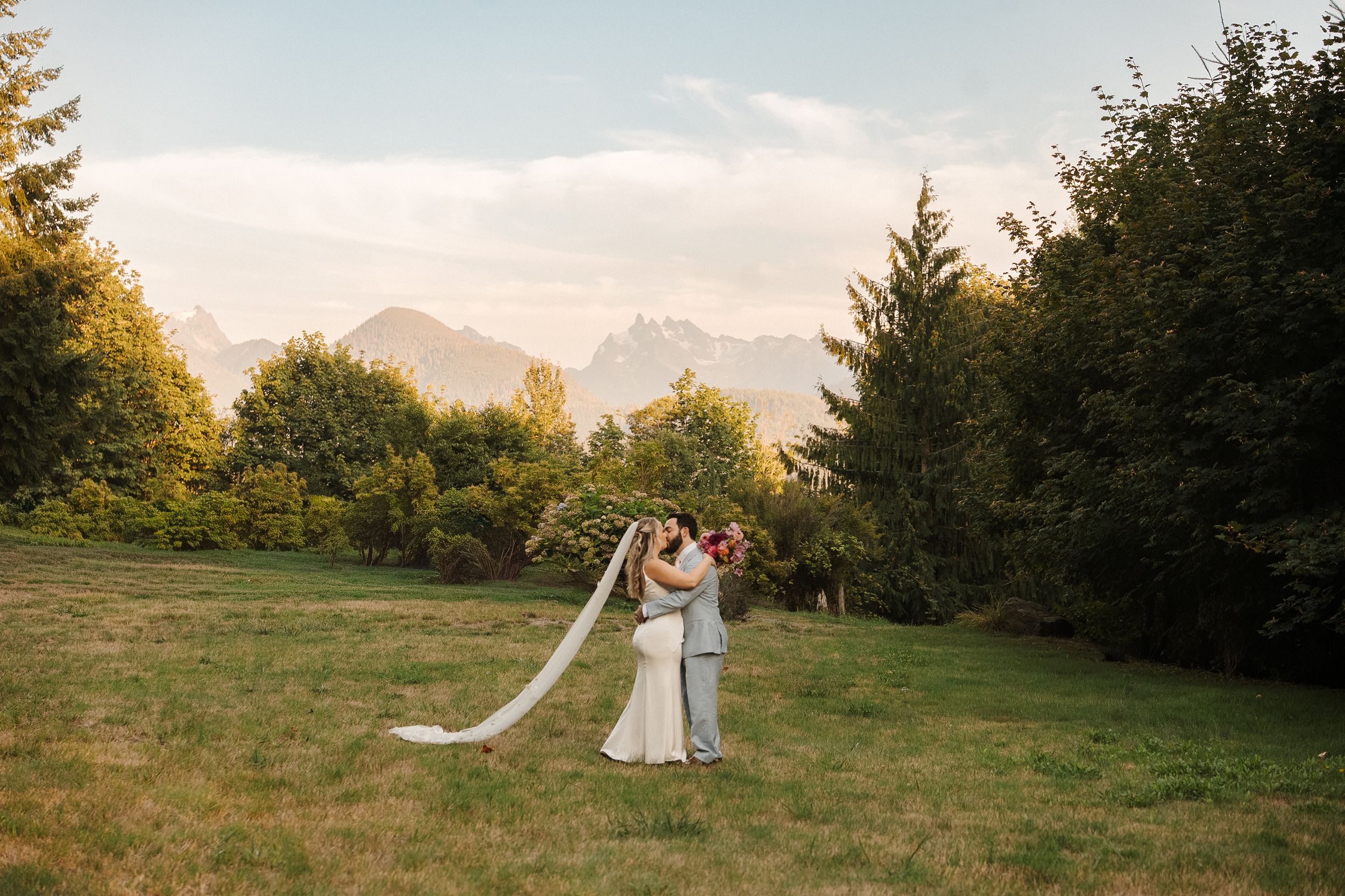 A wedding couple embraces in a golden meadow with majestic mountain peaks and evergreen trees in the background at sunset.
