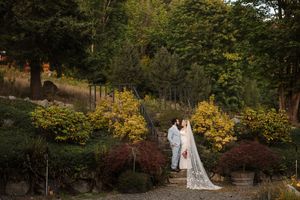 A couple in wedding attire stands together on a hillside garden path surrounded by autumn foliage.
