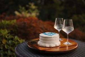 Small white birthday cake with blue fondant decorations sits on a wooden table with wine glasses against autumn foliage backdrop.