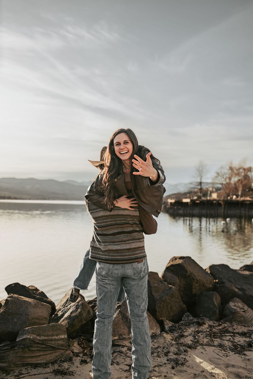 Person laughing joyfully on rocky shoreline with mountains and lake in background.