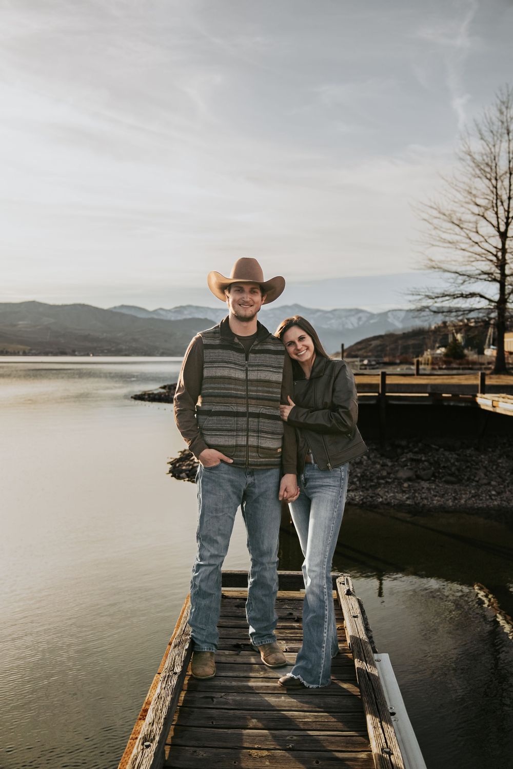 Couple standing together on wooden dock overlooking serene lake and mountains.