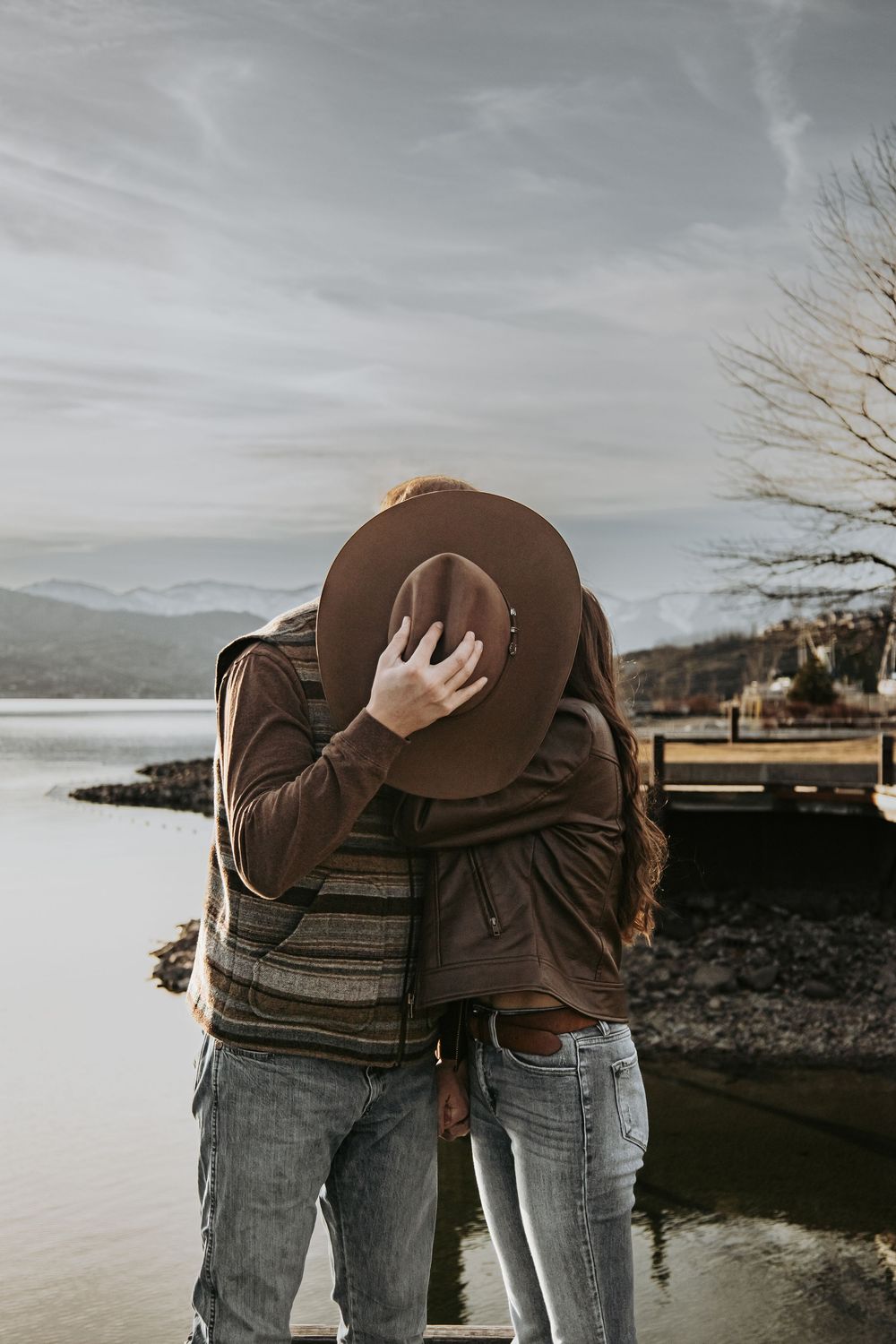 Couple embracing on lakeside dock with mountains and cloudy sky in background.