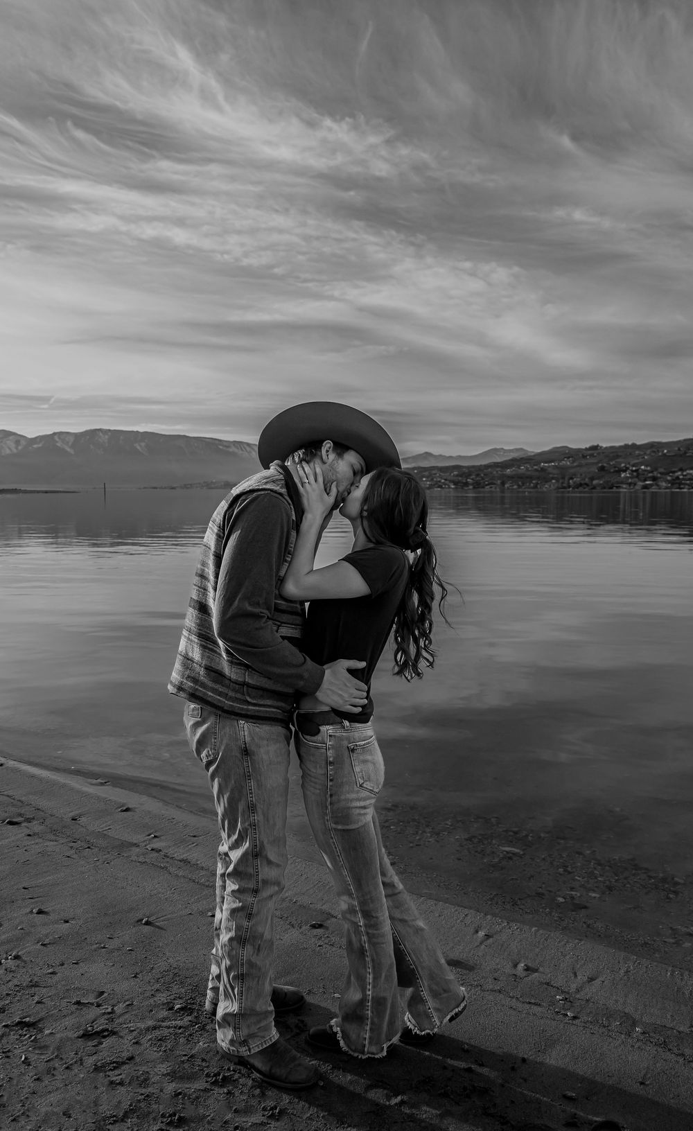 Romantic couple sharing an intimate kiss on a beach with mountains and cloudy skies in the background.