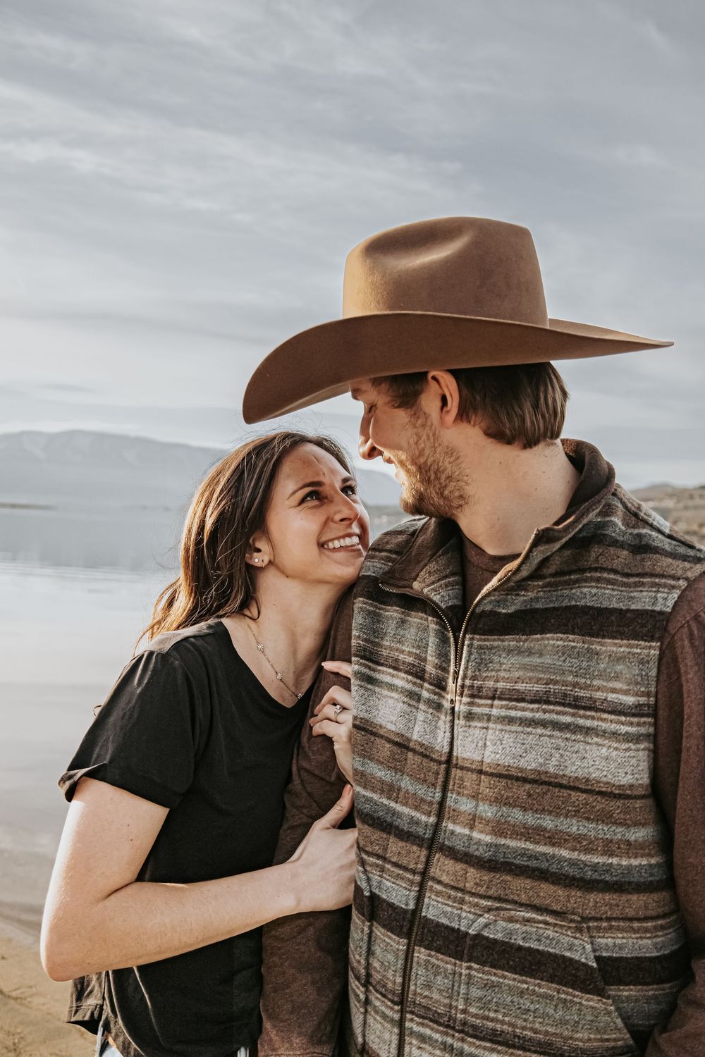 Couple sharing a tender moment on a lakeside beach with mountains in the distance.