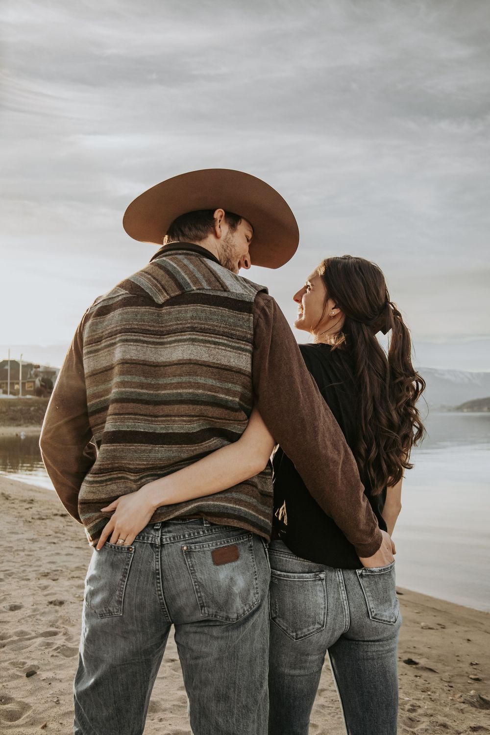 Couple embracing while walking along a scenic beach with mountains and water views.