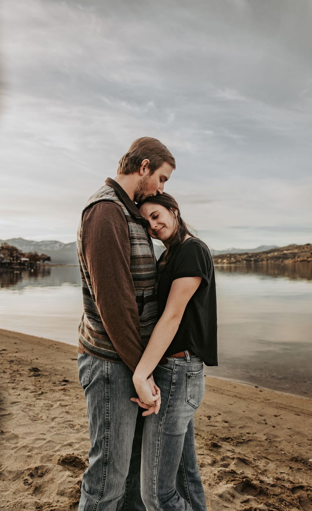 Intimate moment between couple standing on beach with mountains and lake in background.