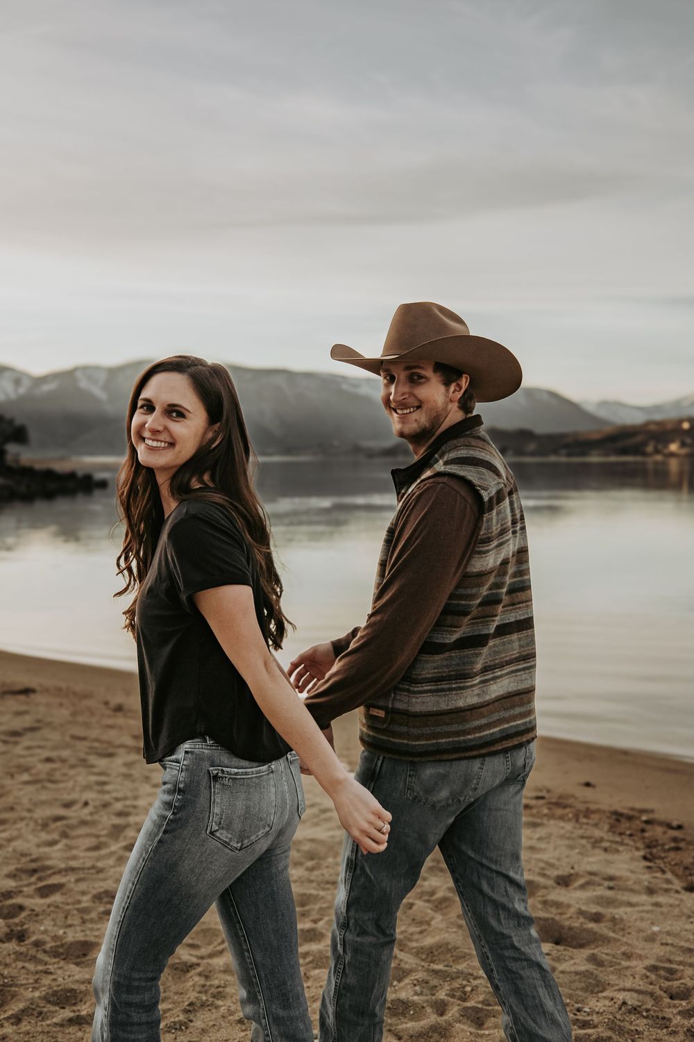 Playful couple walking hand in hand on beach with mountain landscape behind them.