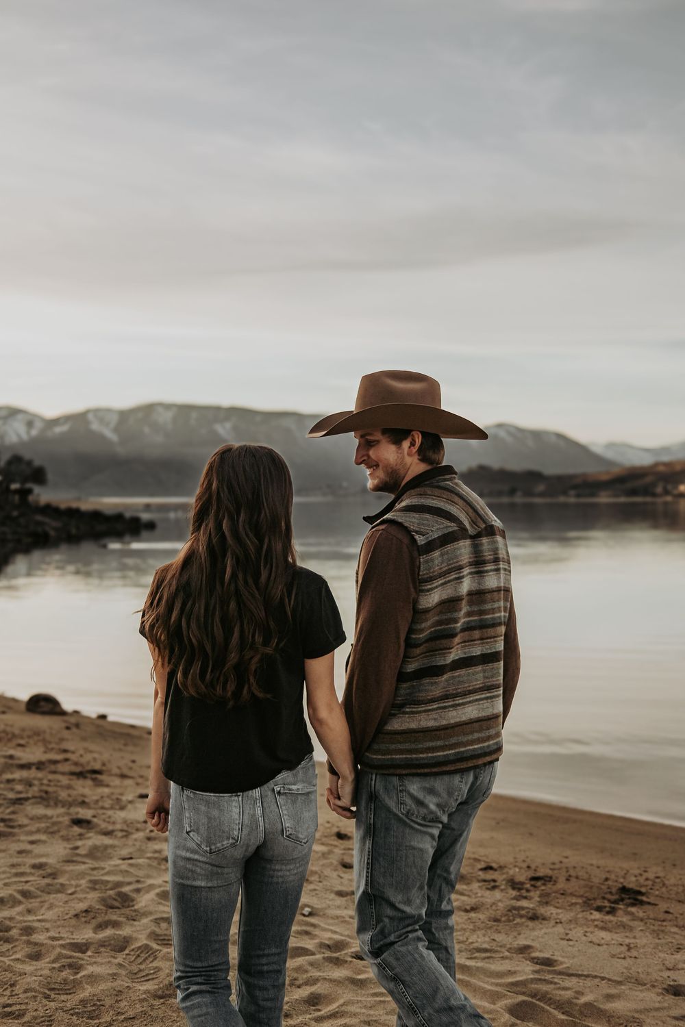 Couple standing together on beach overlooking mountain lake at sunset.