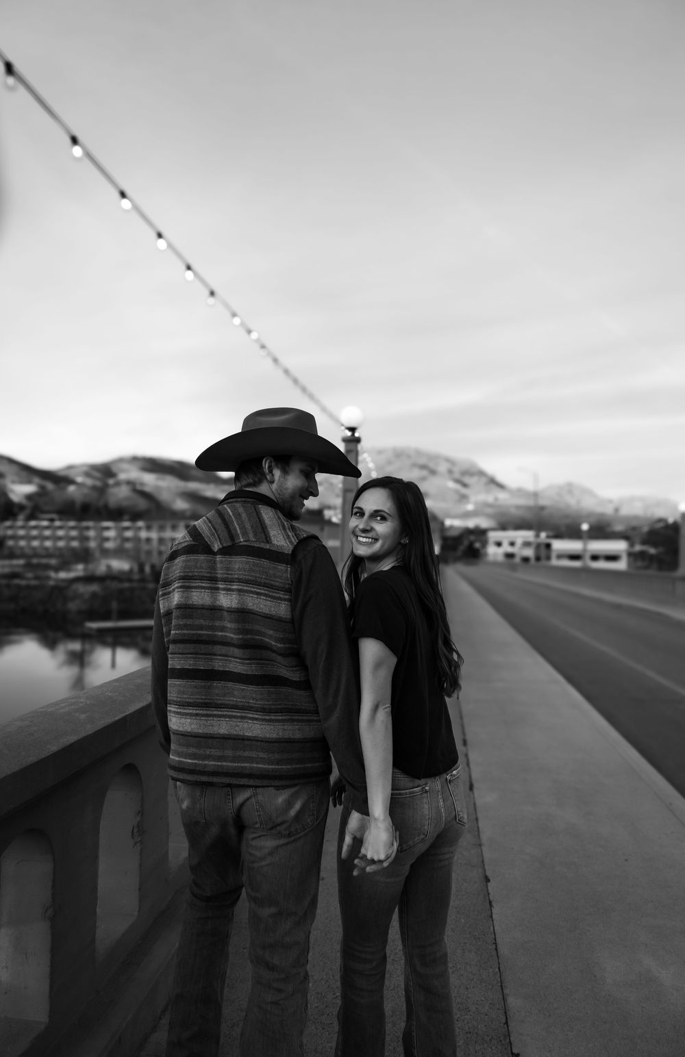 Couple sharing a moment on bridge with string lights and mountains in background.