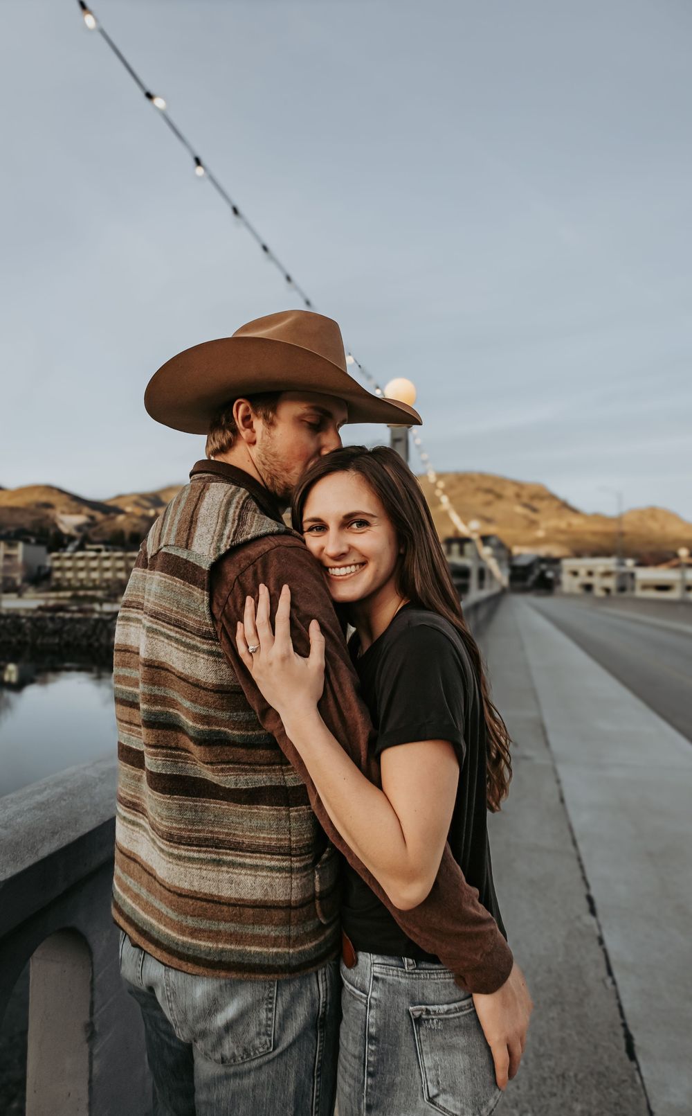 A romantic couple embraces on a rural road wearing western attire against a cloudy sky.