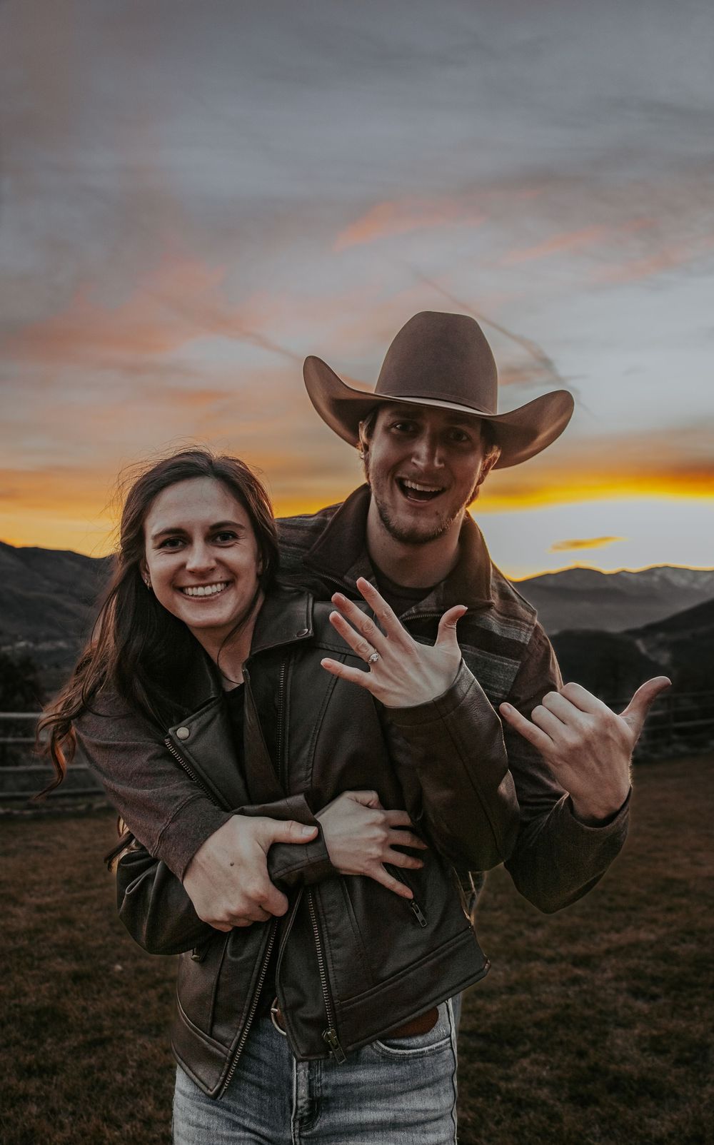 Playful couple in leather jackets making hand gestures against mountain sunset.