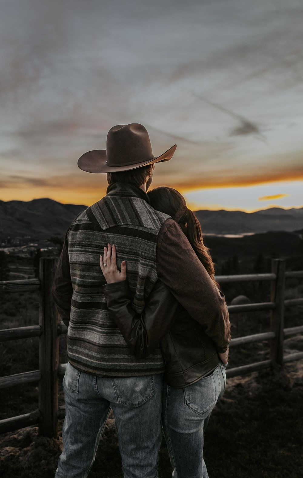 Silhouetted couple embracing by wooden fence during mountain sunset.