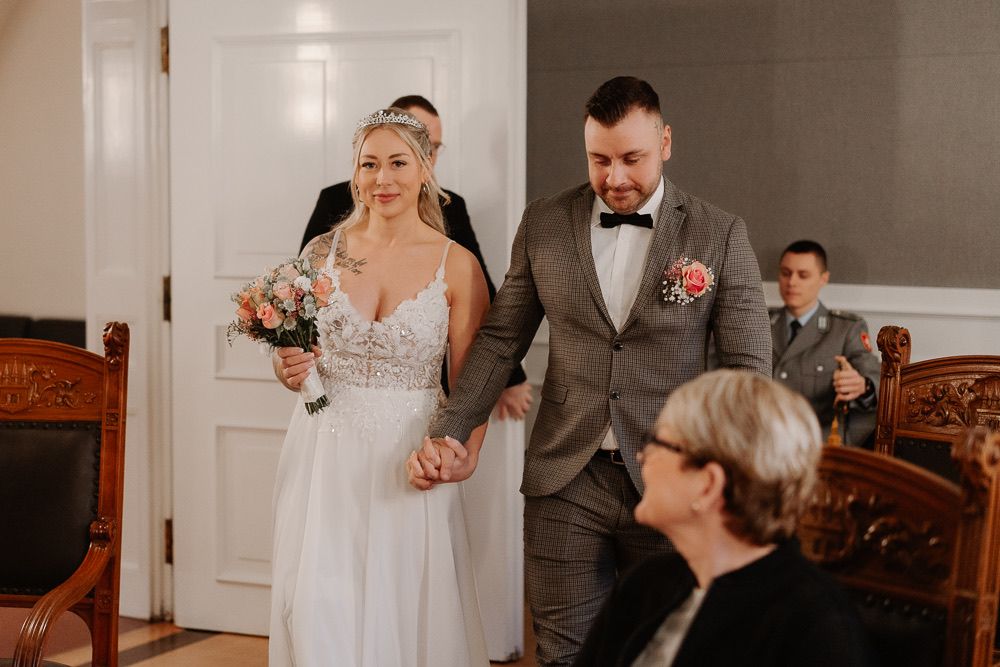 Wedding party members stand together in formal attire against white walls during an indoor ceremony.