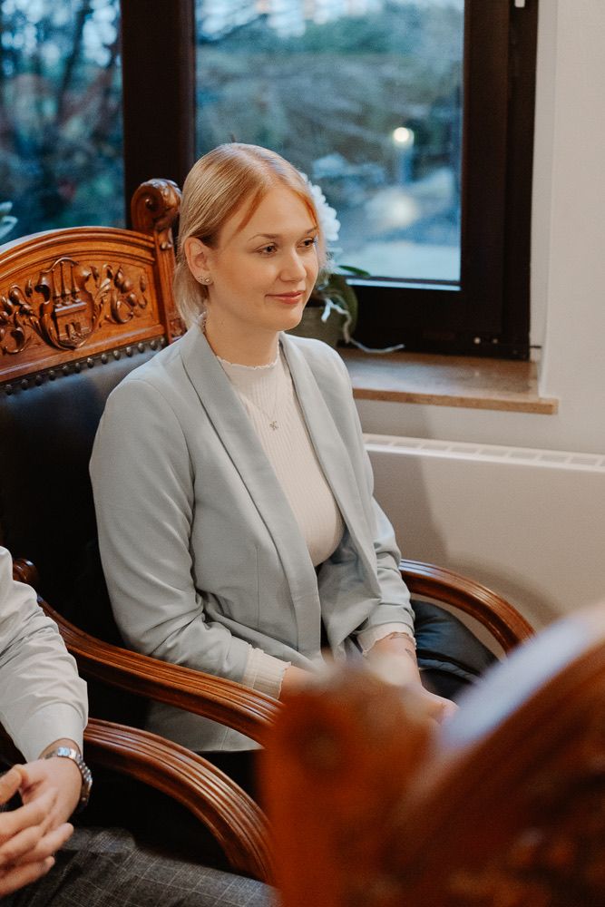 Professional woman in light gray blazer sits thoughtfully by window in elegant wooden chair.