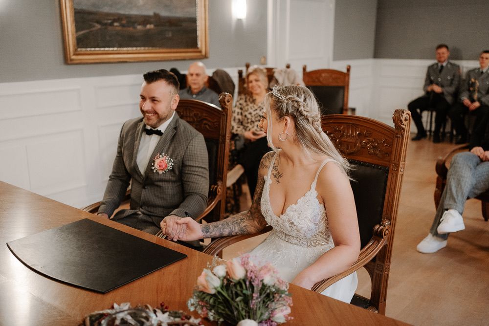 Couple sits at a wooden table during an intimate indoor wedding ceremony with soft natural lighting.
