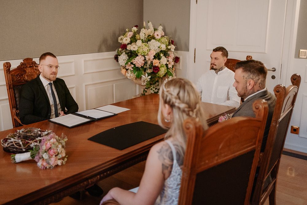 Wedding guests seated in wooden pews during an indoor ceremony with floral arrangements visible.