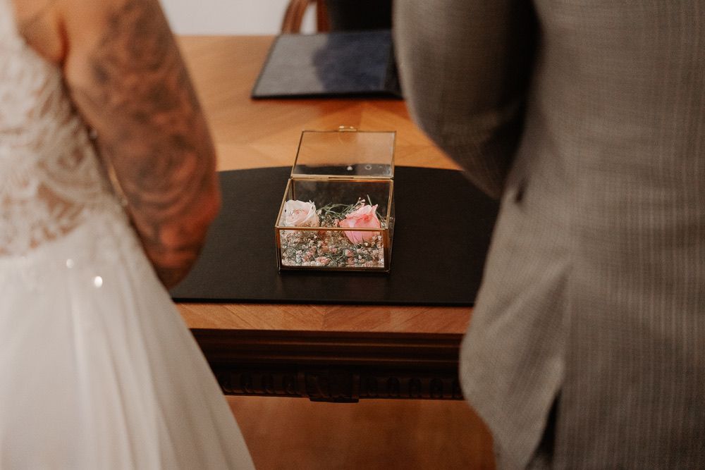 Wedding rings rest in a clear glass box on a wooden surface between a white dress and gray suit.