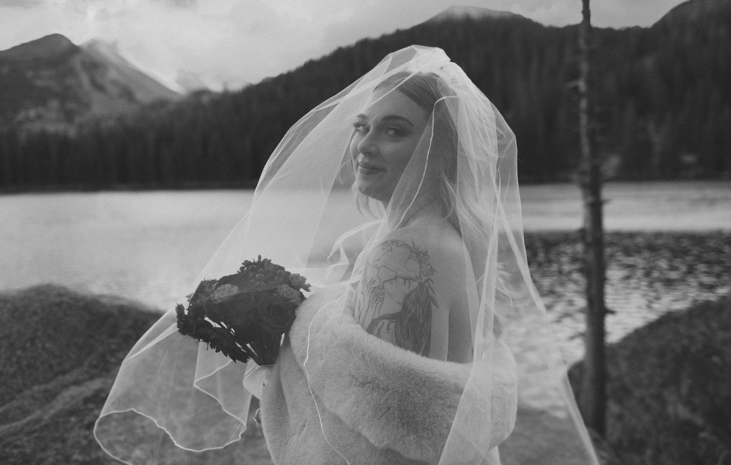 A bride in a flowing white veil and gown stands against a scenic mountain lake backdrop in black and white.