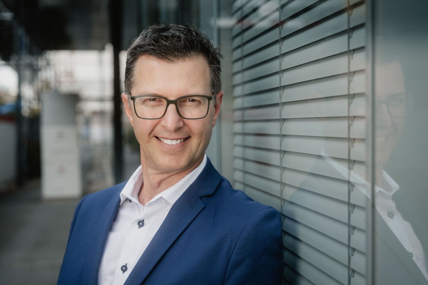 Professional headshot of someone wearing glasses and a navy blue suit jacket against an office building exterior.