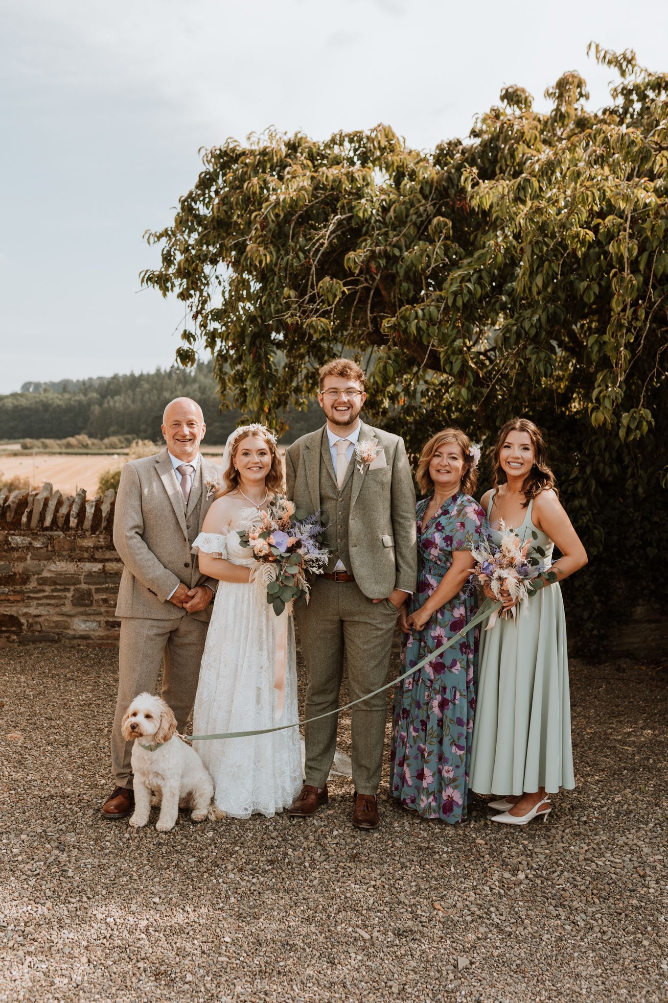 Wedding group photo outdoors with stone wall backdrop and white dog sitting at Wilde Lodge