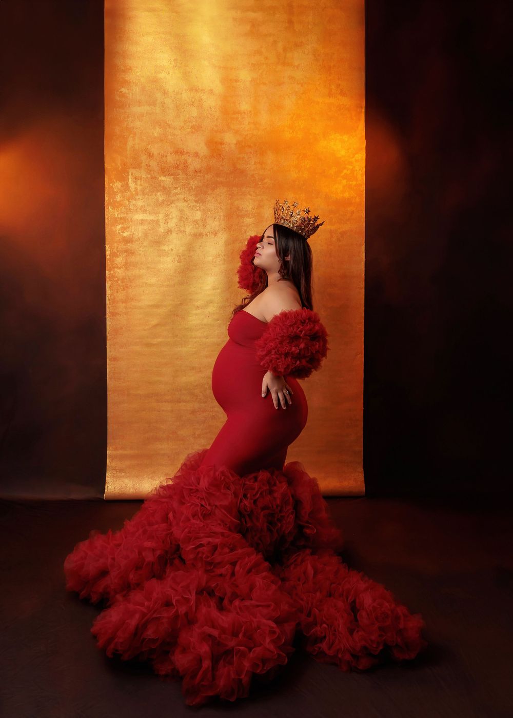 Pregnant woman in red ruffled mermaid gown wearing gold crown poses against golden backdrop.