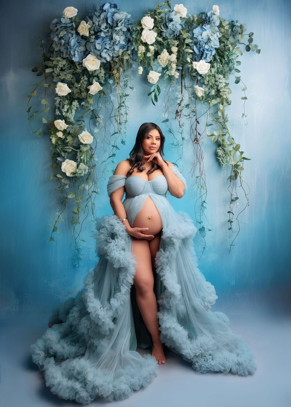 Pregnant woman in elegant light blue ruffled maternity gown posing under blue and white floral arch.