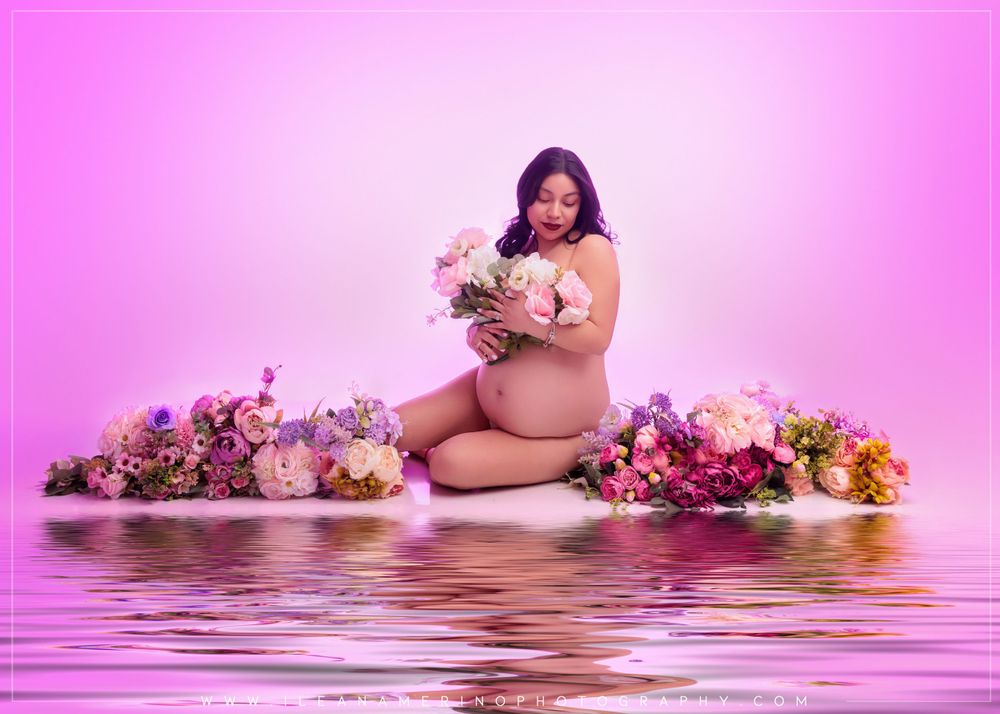 Pregnant woman sits among pink and purple flowers holding bouquet in soft pink-lit maternity studio portrait.