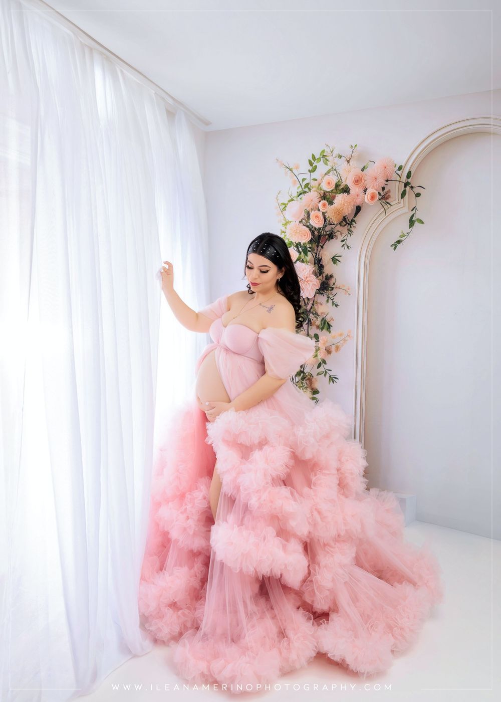 A pregnant woman in a voluminous pink tulle gown poses near a floral arch with roses.