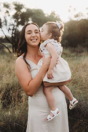 A smiling woman in a white dress holds a toddler girl in a white outfit who kisses her cheek outdoors.