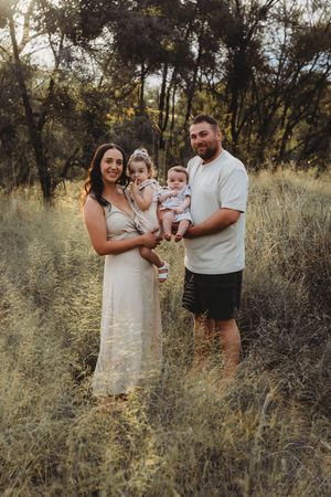 Family of four posing outdoors in golden hour light, parents holding two young children in a lush grassy field.