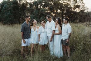 Family of six in white outfits laughing together in a sunlit grassy field surrounded by trees.