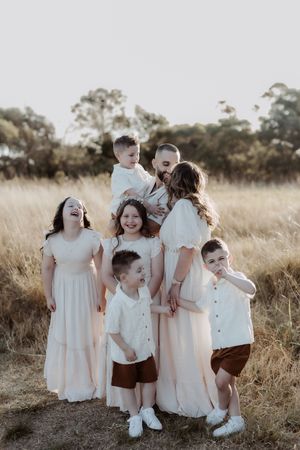 Children and adults in white dresses and brown shorts playing together in a grassy field at sunset.