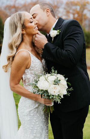 Groom in black suit tenderly kissing bride on cheek as she holds white rose bouquet outdoors.