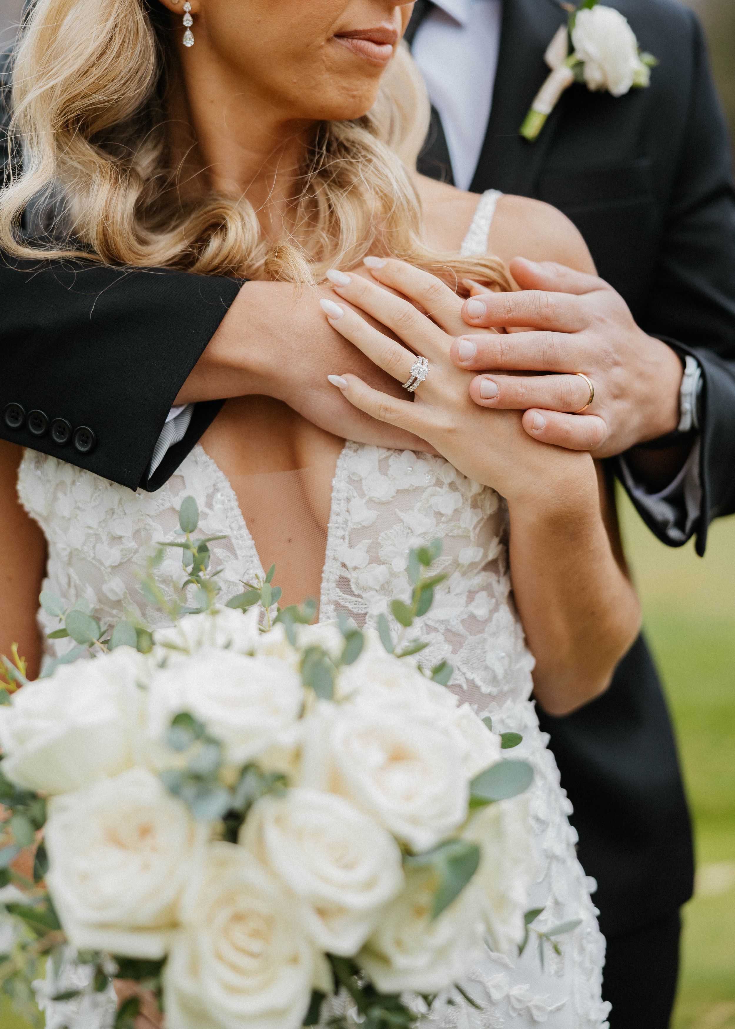 Bride and groom embracing, showcasing wedding rings and a white rose bouquet with eucalyptus.