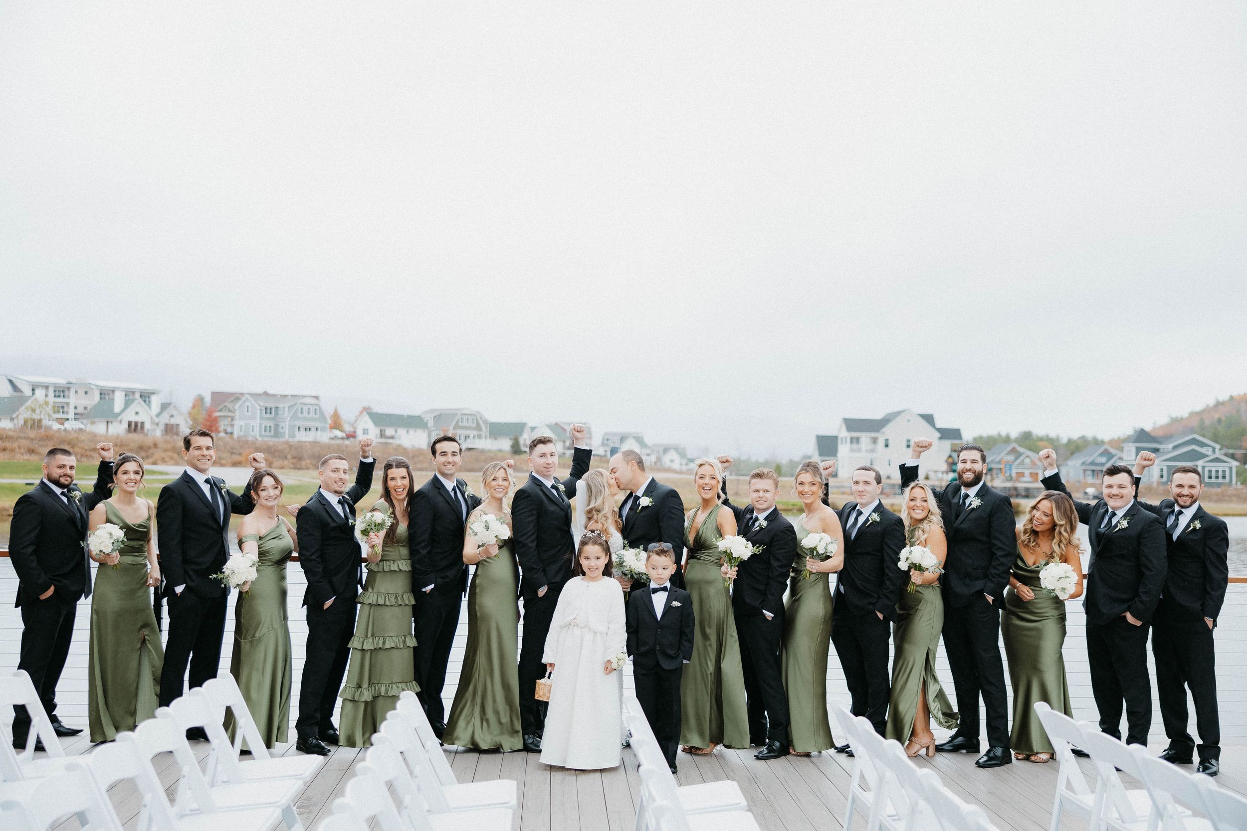 Large wedding party in olive green and black attire posing on an outdoor deck.