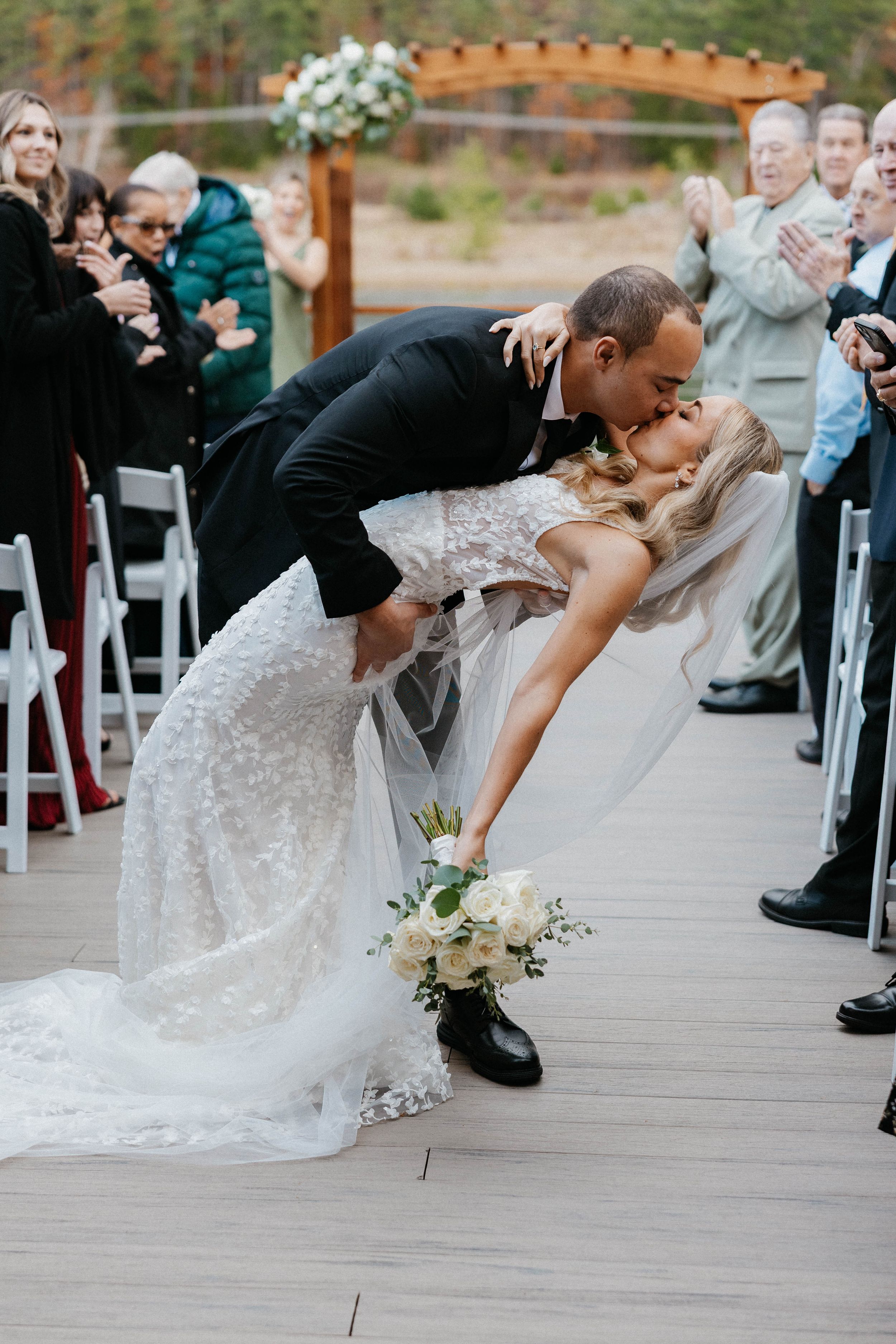 Groom dipping bride for a kiss in the aisle surrounded by applauding wedding guests.